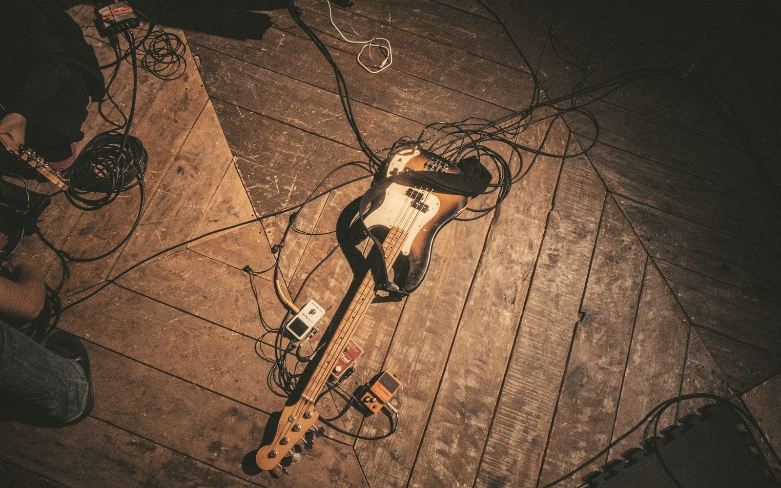 A bass guitar lies on a wooden stage floor amid tangled cables and pedals